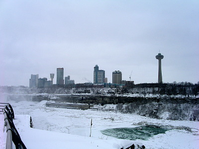 [Overcast skies nearly make the image seem black and white, but there is some hints of blue in what little wter is visible on the ice and snow-covered river. At the top of the hillside are buldings of various shapes and sizes including a tower with a multi-level circular section at the top which I'm sure is used for viewing the Falls.]